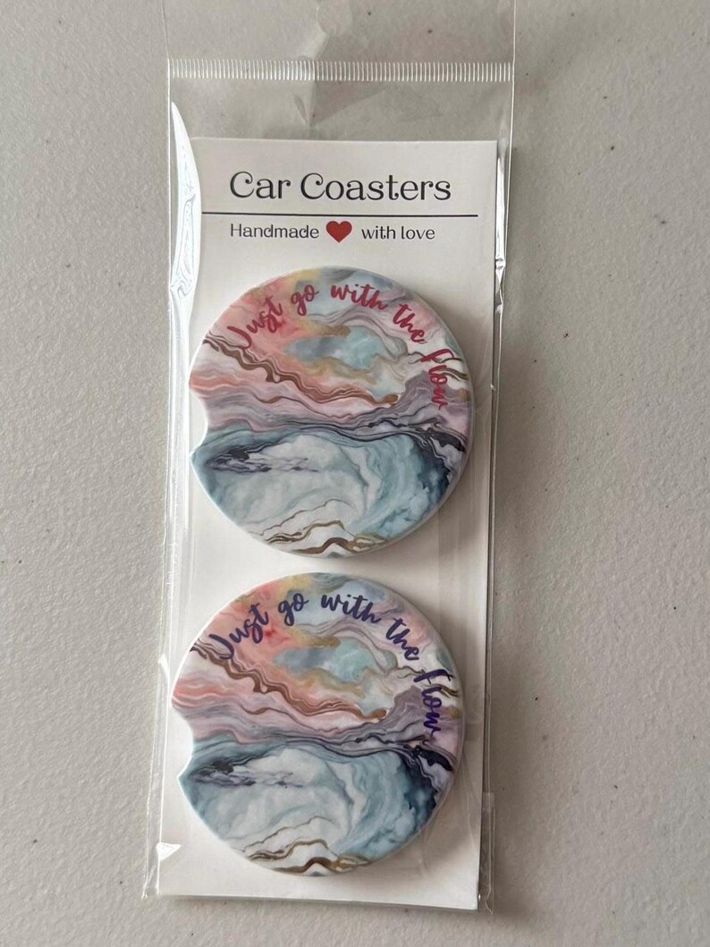 May include: Two car coasters with a colourful abstract design in pink, blue, and gold tones. The coasters have the text "Just go with the flow" in a cursive font. The packaging reads "Car Coasters, Handmade with love."