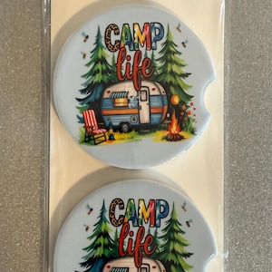 May include: Two car coasters with a light blue background, featuring a camping scene with a camper, trees, and a campfire. The coasters have the words "CAMP life" in colourful letters. The packaging reads "Car Coasters" and "Handmade with love."