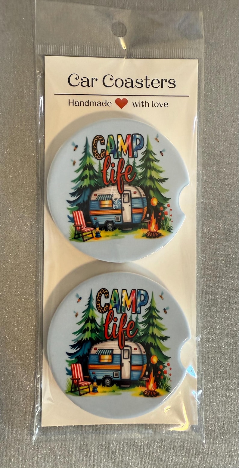May include: Two car coasters with a light blue background, featuring a camping scene with a camper, trees, and a campfire. The coasters have the words "CAMP life" in colourful letters. The packaging reads "Car Coasters" and "Handmade with love."