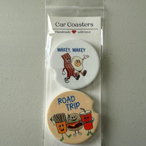 May include: Two car coasters in a clear plastic sleeve. One coaster features a cartoon bacon strip and egg with the text "WAKEY, WAKEY." The other coaster has a cartoon burger, fries, and drink with the text "ROAD TRIP."
