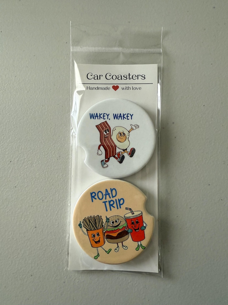 May include: Two car coasters in a clear plastic sleeve. One coaster features a cartoon bacon strip and egg with the text "WAKEY, WAKEY." The other coaster has a cartoon burger, fries, and drink with the text "ROAD TRIP."