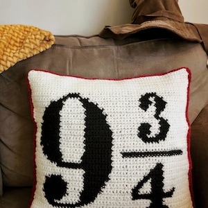 Pode incluir: Uma almofada branca de crochê com borda vermelha apresenta o número "9 3/4" em preto. Um Chapéu Seletor marrom é visível no fundo, sugerindo um tema de fantasia. Um cobertor amarelo texturizado também é visível.