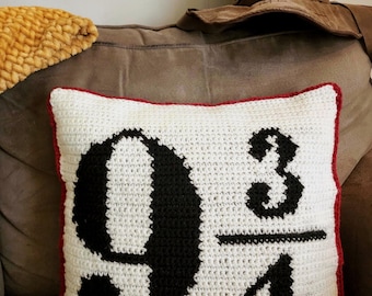 May include: A white crochet pillow with a red border features the number "9 3/4" in black. A brown Sorting Hat is visible in the background, suggesting a theme related to a popular fantasy series. A yellow textured blanket is also visible.