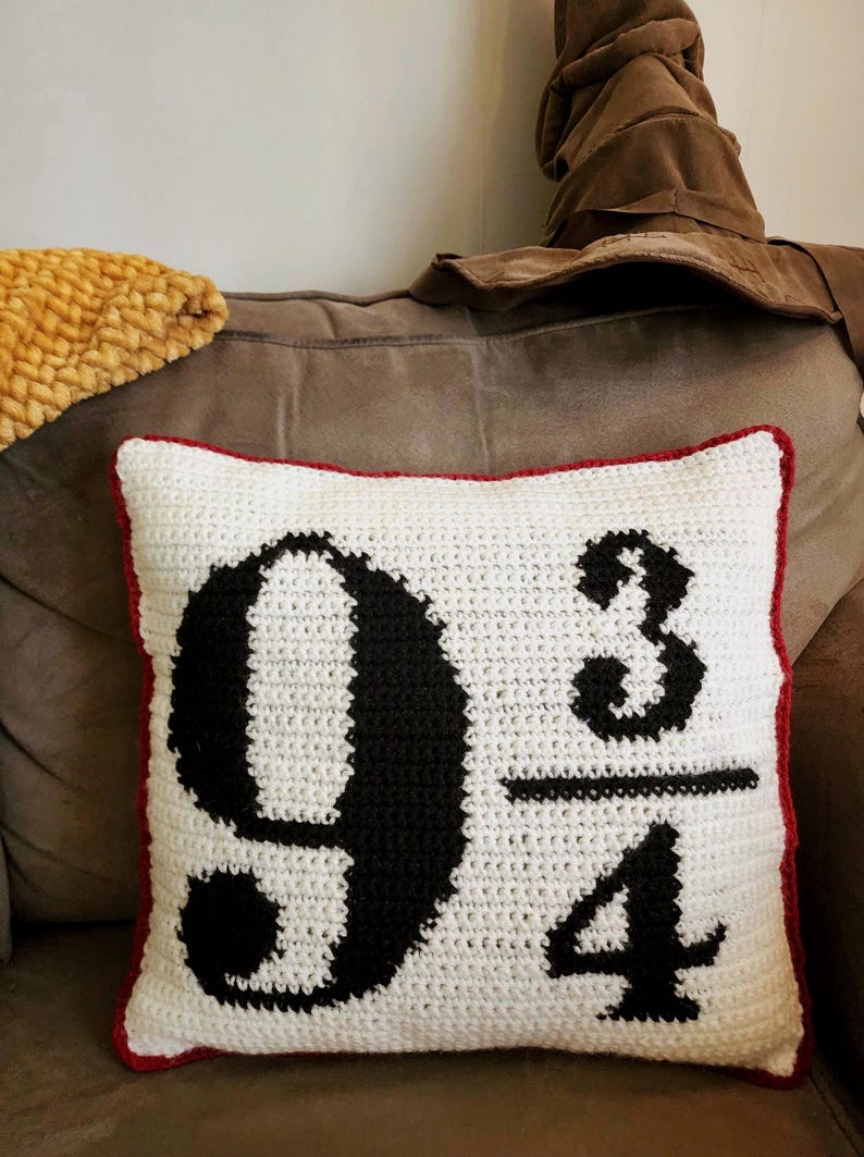 May include: A white crochet pillow with a red border features the number "9 3/4" in black. A brown Sorting Hat is visible in the background, suggesting a theme related to a popular fantasy series. A yellow textured blanket is also visible.