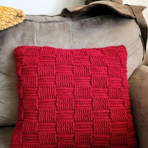 May include: A rectangular red knitted pillow with a woven basket weave pattern. The pillow is resting on a gray couch. A brown hat is behind the pillow.