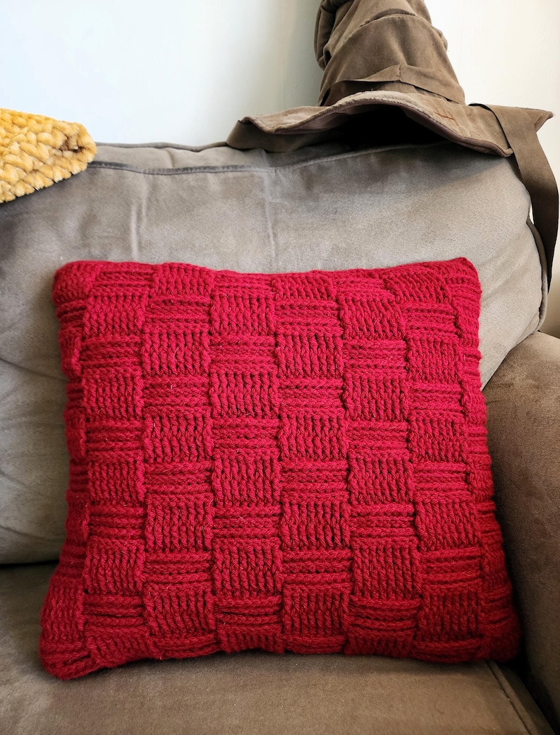 May include: A rectangular red knitted pillow with a woven basket weave pattern. The pillow is resting on a gray couch. A brown hat is behind the pillow.