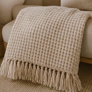 Peut inclure: Une couverture beige en tricot gaufré avec une bordure à franges. La couverture est drapée sur un canapé de couleur claire. Le tissage texturé et la palette de couleurs neutres créent une esthétique chaleureuse et invitante.