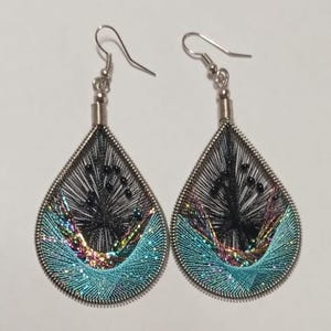 May include: A pair of teardrop-shaped earrings with a silver-tone metal frame. The earrings feature black threadwork at the top, transitioning to a turquoise and iridescent thread design at the bottom. Small black beads are incorporated into the design.