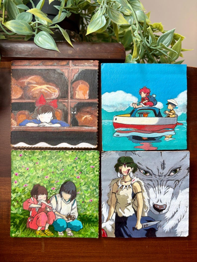 May include: Four square paintings featuring anime characters. One shows a girl in a bakery, another a boy on a boat, a third two children in a field, and the last a woman with a wolf. The paintings are on a wooden surface.