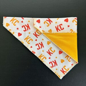 May include: A white dog bandana with a repeating pattern of red and orange "KC" text and heart shapes. The reverse side is solid yellow. The bandana is folded in a triangle shape.
