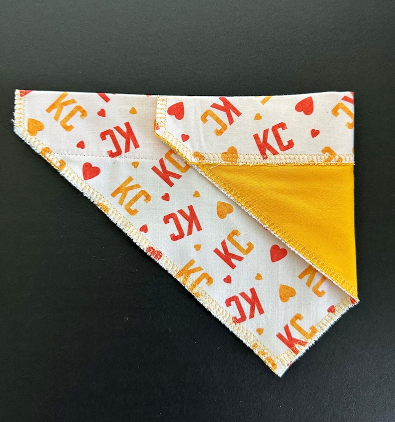 May include: A white dog bandana with a repeating pattern of red and orange "KC" text and heart shapes. The reverse side is solid yellow. The bandana is folded in a triangle shape.