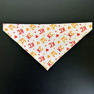May include: White triangular dog bandana with a repeating pattern of red and orange "KC" letters and heart shapes. The edges are finished with a yellow stitch.