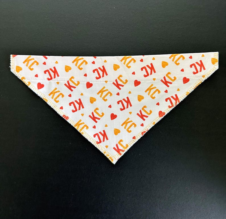 May include: White triangular dog bandana with a repeating pattern of red and orange "KC" letters and heart shapes. The edges are finished with a yellow stitch.