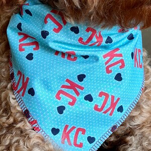 May include: A light blue dog bandana with the letters "KC" in red, interspersed with small black heart shapes. The bandana has a white dotted pattern and a red stitched border. The dog's fur is a light brown color.