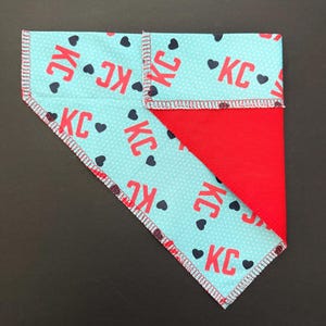 May include: A reversible pet bandana with a light blue pattern featuring red "KC" text and black hearts. The reverse side is solid red. The edges are finished with a white and red zigzag stitch.