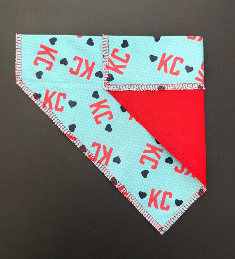 May include: A reversible pet bandana with a light blue pattern featuring red "KC" text and black hearts. The reverse side is solid red. The edges are finished with a white and red zigzag stitch.