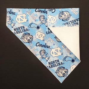 May include: A light blue and white bandana featuring the text "North Carolina" and "UNC" along with Tar Heel logos and graphics. The bandana is folded, revealing a white fabric on the inside.