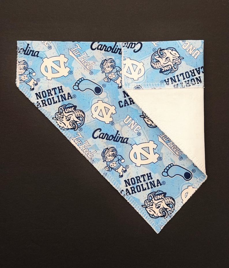 May include: A light blue and white bandana featuring the text "North Carolina" and "UNC" along with Tar Heel logos and graphics. The bandana is folded, revealing a white fabric on the inside.