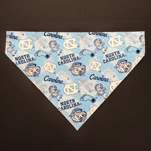 May include: A light blue bandana with a repeating pattern of white UNC logos, Tar Heel text, and cartoon figures. The bandana is triangular in shape and made of fabric.