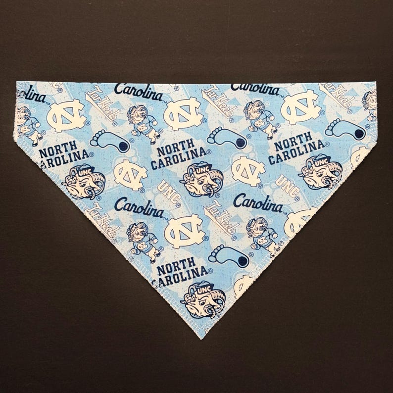 May include: A light blue bandana with a repeating pattern of white UNC logos, Tar Heel text, and cartoon figures. The bandana is triangular in shape and made of fabric.