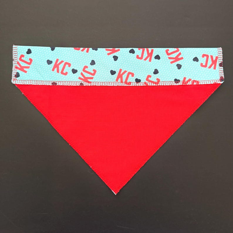 May include: Red and light blue dog bandana. The top section is light blue with the letters "KC" and black hearts. The bottom section is solid red. The bandana is triangular in shape.