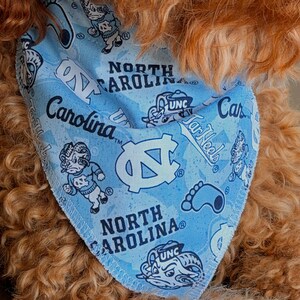May include: Light blue dog bandana featuring the words "North Carolina" and "Carolina" along with the UNC logo and Tar Heels graphics. The bandana is patterned with various UNC mascot illustrations.