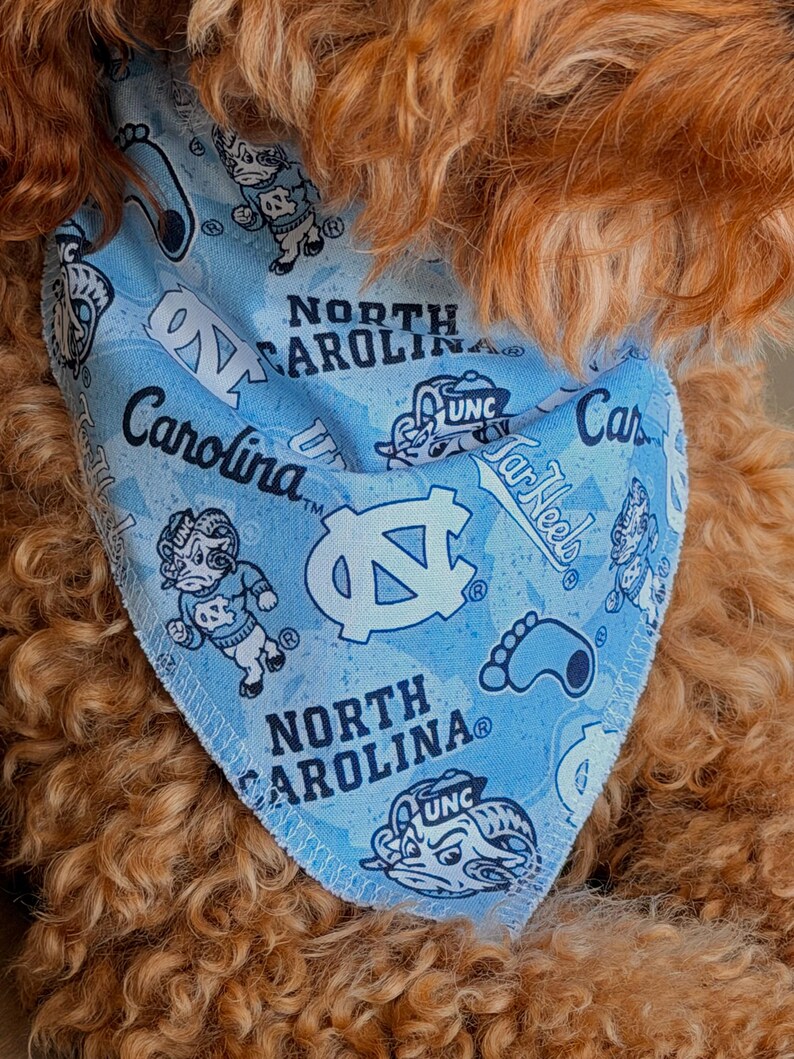 May include: Light blue dog bandana featuring the words "North Carolina" and "Carolina" along with the UNC logo and Tar Heels graphics. The bandana is patterned with various UNC mascot illustrations.