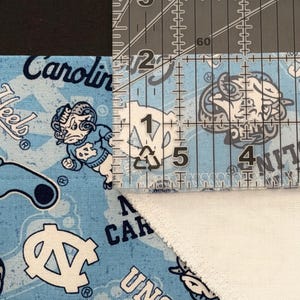 May include: A close-up shot of a light blue fabric with a repeating pattern of the UNC logo and the word "Carolina" in white. A clear ruler is placed over the fabric, showing measurements in inches. The fabric is cut at an angle.