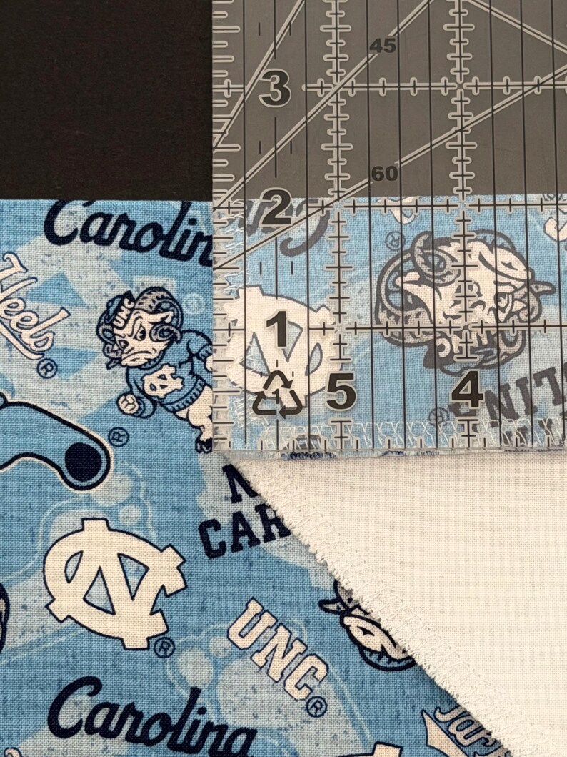 May include: A close-up shot of a light blue fabric with a repeating pattern of the UNC logo and the word "Carolina" in white. A clear ruler is placed over the fabric, showing measurements in inches. The fabric is cut at an angle.