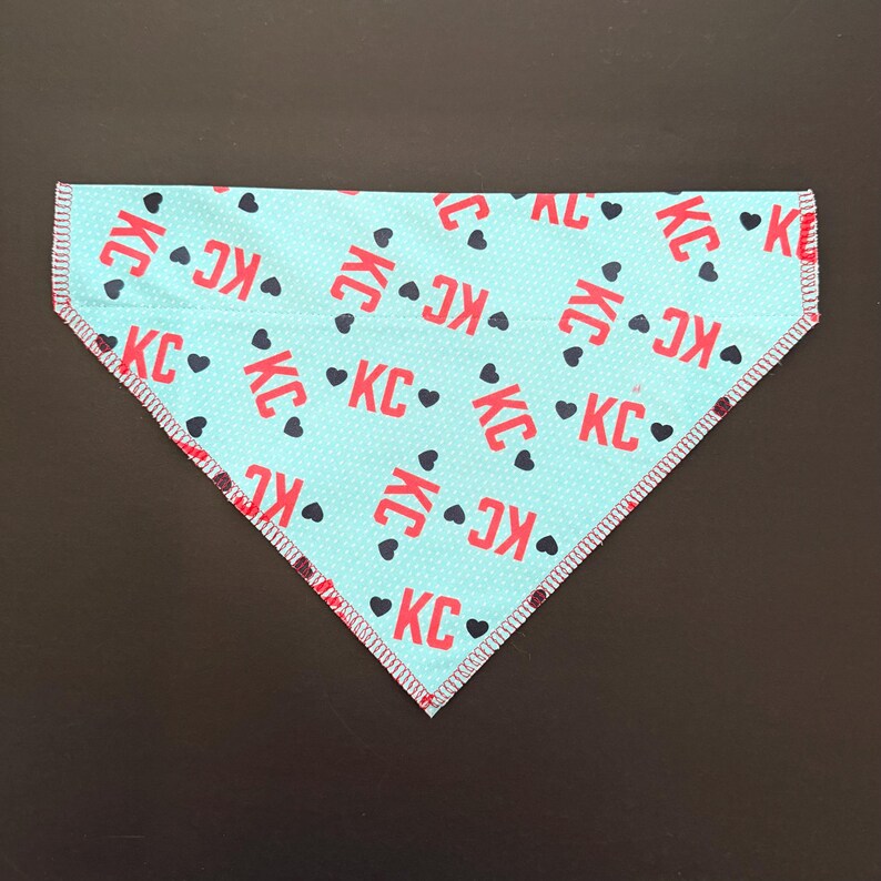 May include: A turquoise dog bandana with a repeating pattern of red "KC" letters and black hearts. The bandana has a polka dot background and a red and white stitched border. The bandana is triangular in shape.