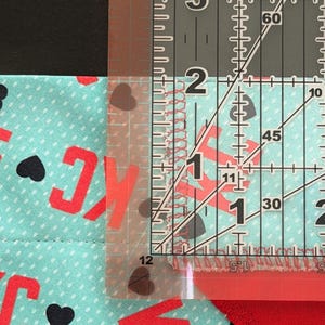 May include: A clear ruler measuring fabric with red letters and black hearts. The fabric is a light blue color with white polka dots. The ruler shows a measurement of approximately 1 inch.