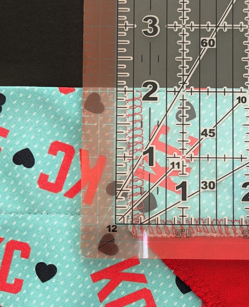 May include: A clear ruler measuring fabric with red letters and black hearts. The fabric is a light blue color with white polka dots. The ruler shows a measurement of approximately 1 inch.