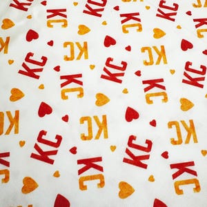 May include: White fabric with a repeating pattern of the letters "KC" in red and orange, interspersed with red and orange heart shapes. The fabric has a slightly wrinkled texture.