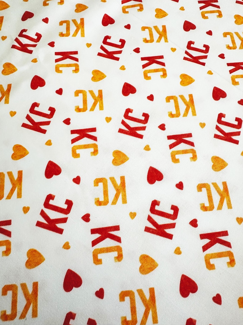 May include: White fabric with a repeating pattern of the letters "KC" in red and orange, interspersed with red and orange heart shapes. The fabric has a slightly wrinkled texture.