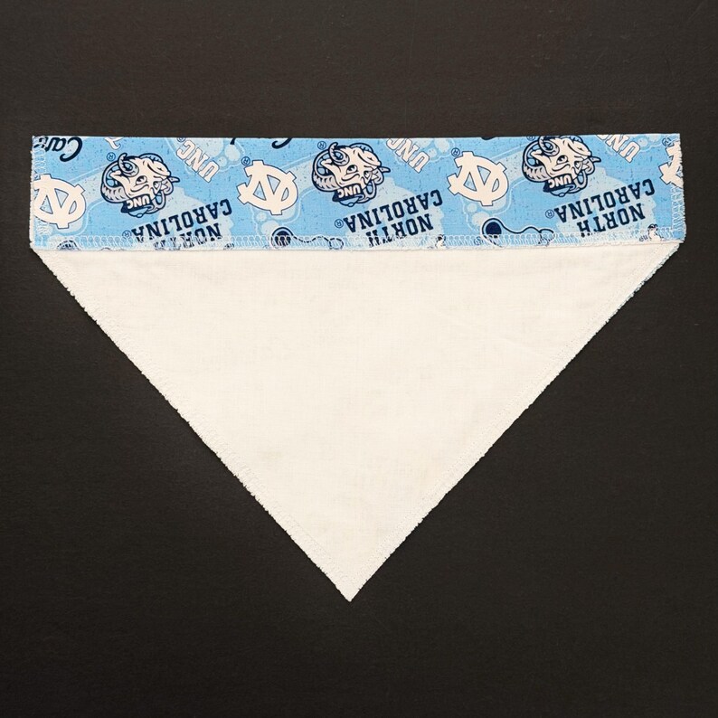 May include: A triangular pet bandana with a light blue top section featuring the words "North Carolina" and the UNC logo in white. The main body of the bandana is white.