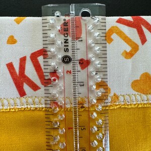 May include: A clear Singer ruler measuring fabric. The fabric is white with orange letters and hearts, and yellow with a stitched edge. The ruler shows measurements in inches.