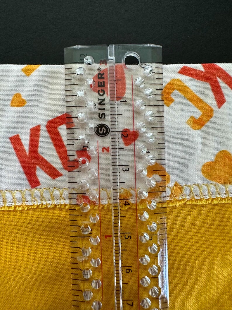 May include: A clear Singer ruler measuring fabric. The fabric is white with orange letters and hearts, and yellow with a stitched edge. The ruler shows measurements in inches.