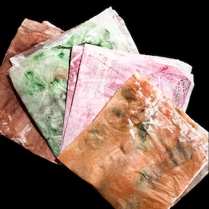 May include: A stack of folded, translucent sheets of paper featuring a marbled, tie-dye effect in various colors including shades of orange, pink, and green. The paper has a crinkled, textured appearance with overlapping layers and irregular patterns.