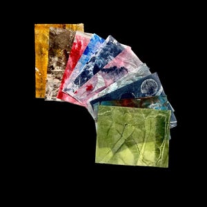 May include: A collection of rectangular paper sheets arranged in a fan shape. Each sheet features a unique abstract pattern with varied textures and colors, including yellow, brown, red, blue, pink, and green, with some showing circular motifs.