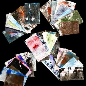 May include: A collection of rectangular paper sheets featuring various abstract paint textures and colors including black, white, blue, red, green, and brown. The papers are arranged in multiple fanned-out stacks, displaying diverse marbled and layered patterns.