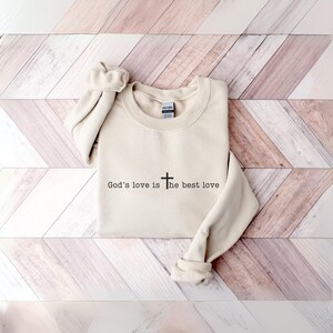 Peut inclure: Un sweat-shirt cr&egrave;me avec le texte "God's love is the best love" en noir, accompagn&eacute; d'une croix. Le sweat-shirt est pli&eacute;, avec les manches retrouss&eacute;es. Le fond est une surface en bois de couleur claire.