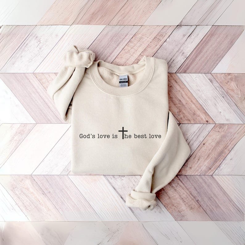 Peut inclure: Un sweat-shirt cr&egrave;me avec le texte "God's love is the best love" en noir, accompagn&eacute; d'une croix. Le sweat-shirt est pli&eacute;, avec les manches retrouss&eacute;es. Le fond est une surface en bois de couleur claire.