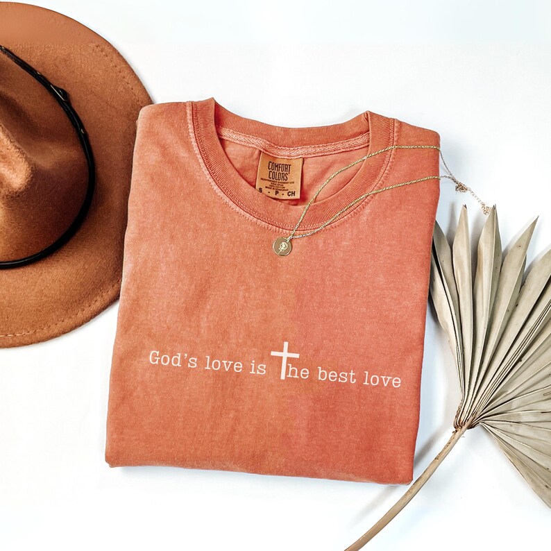 Peut inclure: T-shirt Comfort Colors corail avec le texte "God's love is the best love" en blanc. Un collier dor&eacute; avec un pendentif repose sur le t-shirt. Un chapeau marron et une feuille de palmier s&eacute;ch&eacute;e sont &eacute;galement dans l'image.