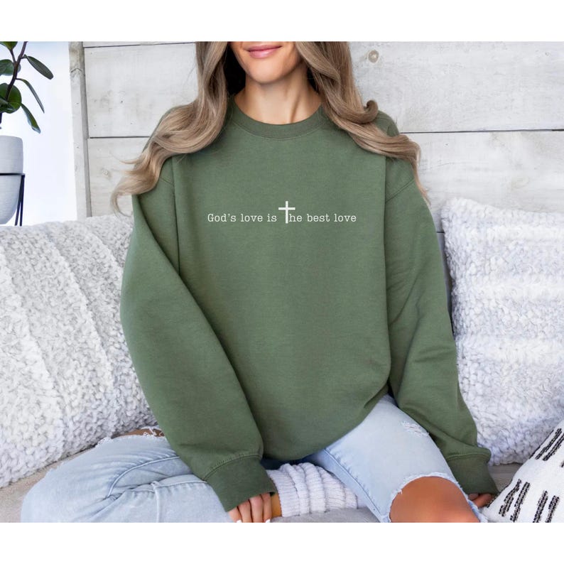 May include: A military green crewneck sweatshirt with the text "God's love is the best love" in white, with a cross symbol wear by a woman sitting on a sofa.