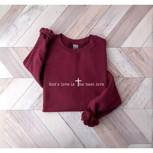 Peut inclure: Un sweat-shirt bordeaux avec l'inscription blanche "God's love is the best love" et une croix. Le v&ecirc;tement est pr&eacute;sent&eacute; sur une surface claire imitation bois.