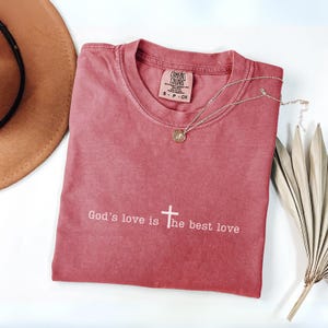 Peut inclure: Un t-shirt rose fan&eacute; avec le texte "God's love is the best love" en blanc. Un collier argent&eacute; avec un pendentif et un chapeau marron sont &eacute;galement visibles. Le t-shirt a un col rond et une petite &eacute;tiquette.