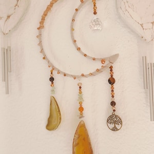 Puede incluir: Adorno de pared artesanal con forma de luna con una media luna clara y texturizada. Cuenta con un cristal, cuentas y rodajas de ágata en tonos marrón, naranja y verde. Un dije metálico del árbol de la vida cuelga debajo.