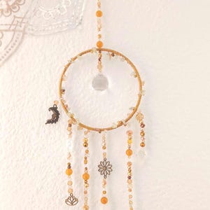Puede incluir: Un atrapasueños artesanal con un aro dorado, adornado con cuentas ámbar y transparentes, y dijes decorativos. Los dijes incluyen una luna creciente, una flor de loto, un sol y un árbol de la vida. Diseñado para colgar en la pared.