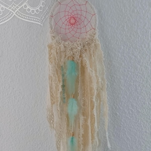 Puede incluir: Un atrapasueños hecho a mano con una red de encaje color crema y un centro rosa. Cuenta con tres plumas azul claro y una cadena de cuentas. El atrapasueños cuelga contra una pared blanca.