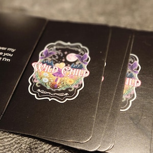 May include: A deck of black cards with the words "I will not lower my voice because you don't like what I'm saying" printed on one card. The other cards have a "WILD CHILD" design with a floral and moon graphic.